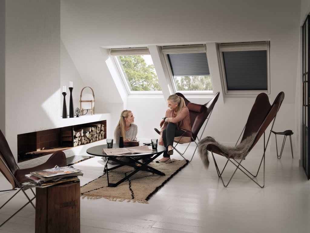Artim Menuisier Cholet VELUX 121234 01 Xxl coin salon aménagé sous les combles avec fauteuils et deux personnes qui sont en train de discuter. Il y a 3 VELUX cote à cote ave des stores,, l'un ouvert, l'autre semi ouvert et le dernier fermé