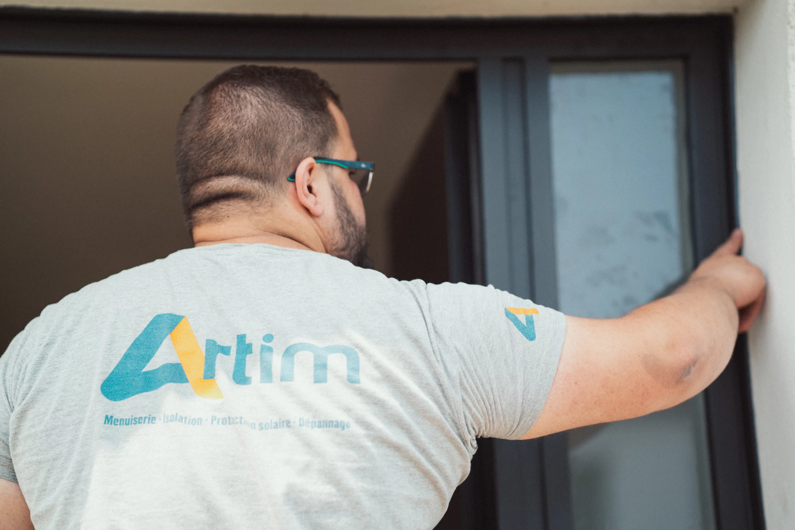 menuisier de dos avec le logo ARTIM sur son tshirt en train de faire un joint sur une fenêtre