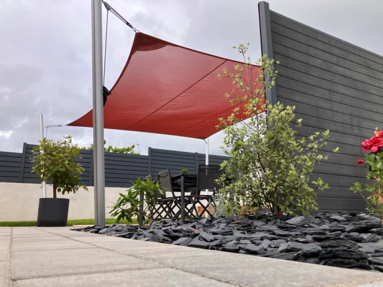 Voile d'ombrage rouge au dessus d'une terrasse avec des mats, vue du dessous faisant apparaitre l'allé et un massif avec de l'ardoise