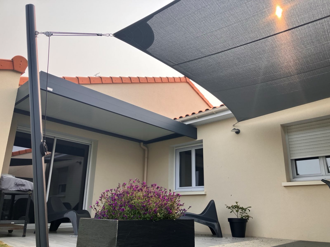 Artim Menuisier Cholet Voile Ombrage Signal 2025 06 03 180824 009 vue sur une terrasse de maison à cholet avec un carport grise et un voile d'ombrage rectangle grise avec petite coin salon et coin terrasse avec table à manger sur la terrasse
