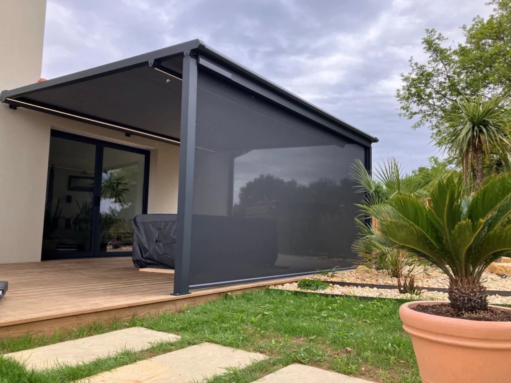 pergola avec store zip sur le devant couleur gris anthracite sur une terrasse bois dans un jardin arboré avec gazon vert et pas japonnais