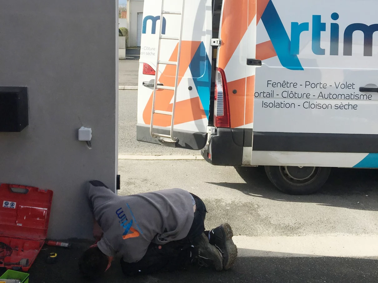 menuisier de chez ARTIM accroupi en traine de réparer un portail de cloture avec vue sur les outils et le camion