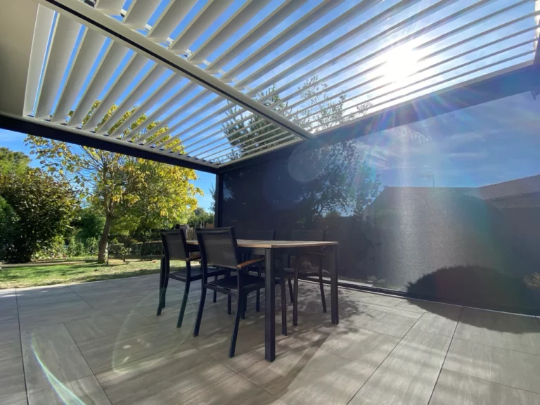 pergola bioclimatique avec 1 store baissé sur une terrasse bois avec une table et chaises et lames ouvertes cholet
