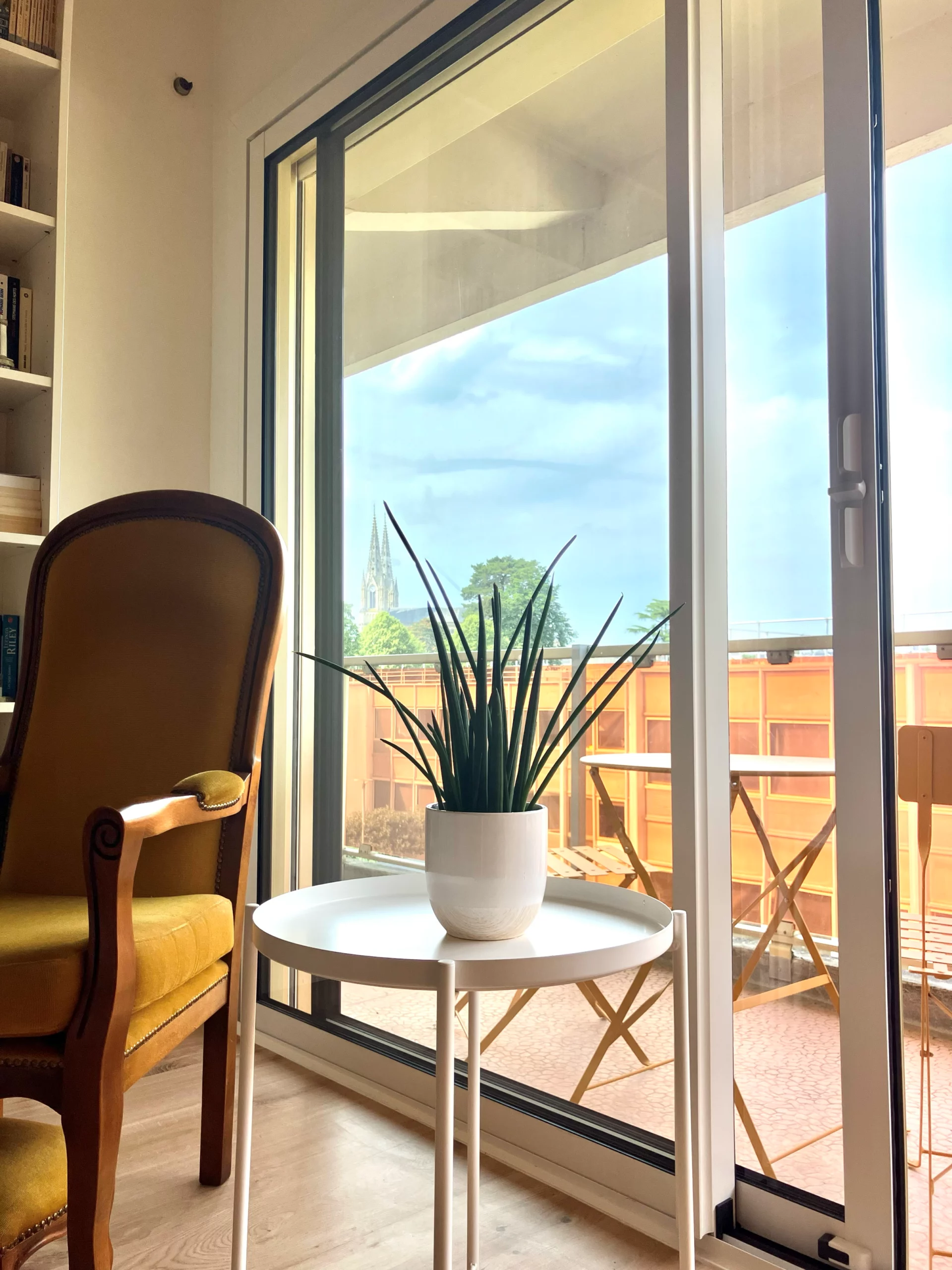 Coin repas lumineux avec porte-fenêtre blanche ouverte sur balcon, vu depuis une table en bois avec plante fleurie au premier plan à Cholet.