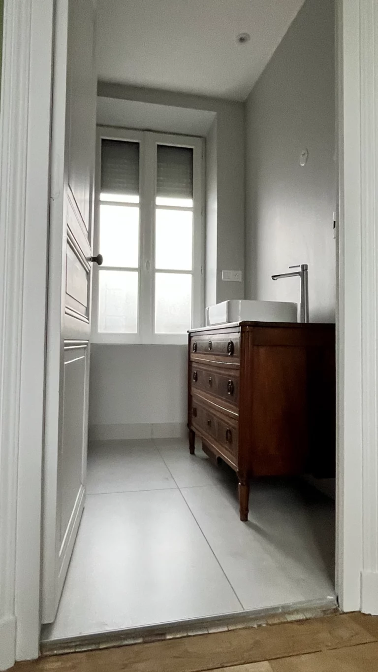 Fenêtre blanche haute dans une salle de bain lumineuse, posée au-dessus d’un meuble ancien dans une maison à Cholet.